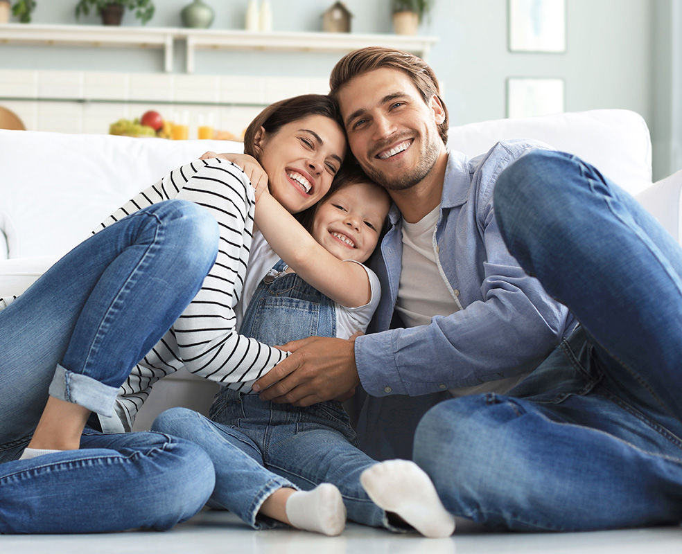 A family of three, including a man and woman with their child, sitting on a couch, smiling and embracing each other.