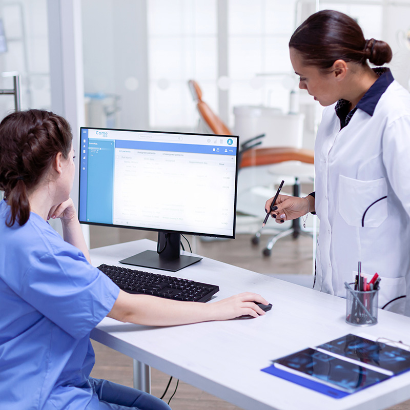 The image depicts a professional setting with a woman in scrubs standing at a desk, interacting with a computer screen, while a man in a lab coat is seated and appears to be observing or assisting her.